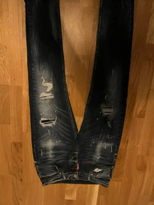 Mörkblå jeans från Dsquared2, stl 33 - Säljer ett par mörkblå jeans från Dsquared2 i storlek 33. Byxorna har slitna detaljer, hål och färgstänk som ger en cool och edgy look. Klassisk femficksmodell med knappgylf och låg midja. Materialet är bomull med lite stretch för skön passform.