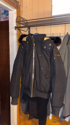 Svart Moose Knuckles - Säljer en svart dunjacka från Moose Knuckles med guldfärgade detaljer och dragkedja. Jackan har svart päls på huvan, flera fickor med tryckknappar och snyggt mönstrat guldigt innerfoder. Perfekt för kalla dagar och riktigt stilren look.