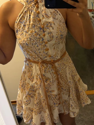 Mönstrad kortklänning från Zara - Säljer en superfin kortklänning från Zara med beige botten och blommigt mönster i blått och gult. Klänningen har hög hals, knappar framtill och ett flätat bälte i midjan. Materialet är lätt och känns som bomull, perfekt för varma dagar.