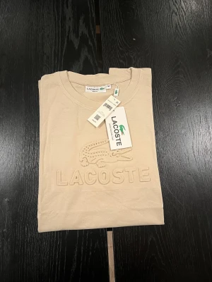 Beige slim fit t-shirt från Lacoste - Snygg beige t-shirt från Lacoste i slim fit-modell. T-shirten har korta ärmar och rund hals, med ett upphöjt Lacoste-logotyptryck framtill. Materialet är mjuk bomull med lite spandex för extra stretch och komfort. Perfekt för dig som gillar stilrena och klassiska plagg.