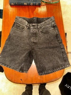 Svarta denimshorts från Carhartt - Säljer ett par svarta denimshorts från Carhartt i modellen 'Newel Short'. De har en klassisk femficksdesign, raka ben och en snygg tvättad look. Materialet är 100% bomull och bakfickan har Carhartt-loggan. Perfekta för en chill och avslappnad stil.