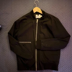 Svart bomberjacka från Topman - Svart bomberjacka från Topman med silverfärgade dragkedjor framtill och på fickorna. Jackan har en klassisk rak passform, ribbade muddar vid ärmslut och nederkant samt långärmad design. Materialet är troligtvis polyester och bomull, vilket gör den lätt och bekväm.