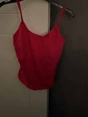 Röd linnetopp med smala axelband - Säljer en enkel och snygg röd linnetopp med smala axelband och v-ringad hals. Toppen har en figurnära passform och är perfekt för varma dagar. Materialet känns mjukt och lätt mot huden. Passar dig som gillar stilrena och färgstarka plagg.