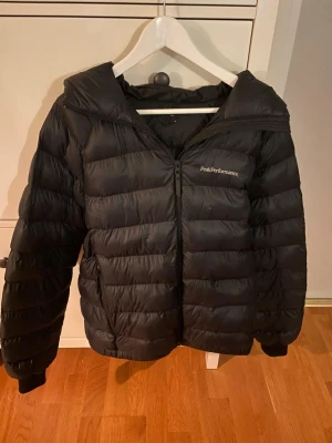 Svart Peak Performance dunjacka M - Svart dunjacka från Peak Performance med huva och dragkedja framtill. Jackan har quiltad design och är tillverkad i syntetmaterial med dunfyllning. Perfekt för kalla dagar och har en clean look med diskret logga på bröstet.