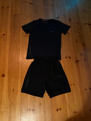 Svarta träningskläder från Under Armour - Säljer ett set med svart t-shirt och svarta shorts från Under Armour. T-shirten har rund hals och ett färgglatt logotryck framtill. Shortsen har elastisk midja och tryckt logga på benet. Perfekt för dig som gillar sportig stil och bekväma material.