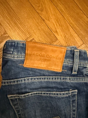 Jacob Cohen Jeans, BARD stl 33 - Säljer ett par klassiska blå jeans från Jacob Cohën Milano. Jeansen har raka ben, fem fickor och snygga kontrastsömmar. Skinnpatch med logga bak i midjan ger en exklusiv touch. Perfekta för dig som gillar stilrena och tidlösa jeans.