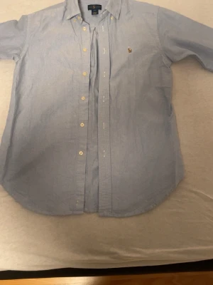 Blå skjorta från Ralph Lauren - Snygg ljusblå skjorta från Ralph Lauren med klassisk krage och broderad logga på bröstet. Skjortan har knappar framtill och är långärmad. Materialet är mjuk bomull som känns skönt mot huden. Perfekt för dig som gillar stilrena och tidlösa plagg.