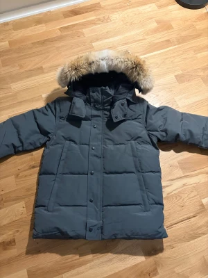 Mörkgrå Canada Goose dunjacka med päls - Säljer en mörkgrå dunjacka från Canada Goose med stor huva och snygg pälsdetalj. Jackan har quiltad design, knappar och dragkedja framtill samt två stora fickor. Perfekt för kalla vinterdagar och riktigt stilren look. Storlek S & M finns i båda svart och grå 