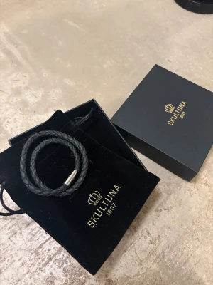 Ett skultuna armband  - Bilderna visar ett svart flätat armband från Skultuna med metallspänne, samt tillhörande ask och tygpåse. Perfekt för någon som vill sticka ut ännu mera med sin outfit! Ny pris ligger runt 900-1200kr 