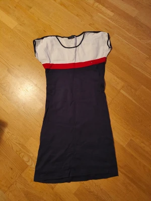 Tommy Hilfiger marinblå kortklänning - Snygg kortklänning från Tommy Hilfiger med klassisk färgblock-design i marinblått, vitt och rött. Klänningen har en enkel, rak form och är tillverkad i mjuk bomull. Detaljerad med Hilfiger-logga på bröstet och korta ärmar. Perfekt för en fräsch och sportig look.