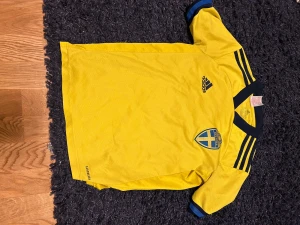 Svensk landslagströja Adidas  - Gul landslagströja från Adidas med blå detaljer och Sverige-emblem på bröstet. Tröjan har klassiska Adidas-ränder på axlarna och Aeroready-material som andas. Perfekt för fotbollsfans som vill representera Sverige. Passar för dig mellan 150-155cm lång