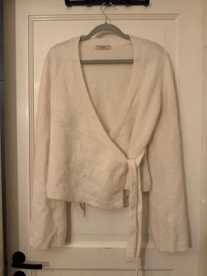 Vit omlottkofta från Gina Tricot - Mysig vit omlottkofta från Gina Tricot i storlek M. Koftan har långa ärmar, ribbade muddar och knytband i midjan. Tillverkad i ett mjukt stickat material som känns skönt mot huden. 