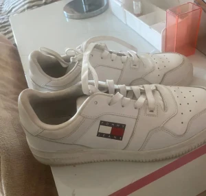 Vita sneakers från Tommy Jeans - Säljer ett par fräscha vita sneakers från Tommy Jeans med klassisk logga på sidan. Skorna har snörning, perforerad tå och platt sula. Tillverkade i skinn med bekväm innersula och rund tå. Perfekta för dig som gillar stilrena och tidlösa sneakers.