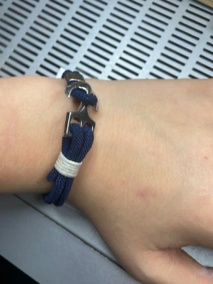 Marinblå armband med ankare - Snyggt marinblått armband med ett silverfärgat ankare som lås. Armbandet är gjort av flätad syntet och har en vit detalj runt banden. Perfekt accessoar för dig som gillar nautisk stil och vill ha något unikt på handleden. väldigt nice och fint att ha till sommaren