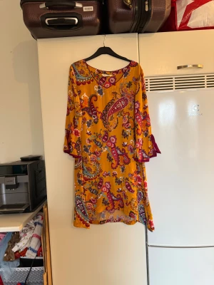 Färgglad kortklänning med paisleymönster - Säljer en superfin kortklänning med paisley och blommigt mönster i starka färger som gul, lila, blå och röd. Klänningen har v-ringning och trumpetärmar med lila kant. Materialet är mjukt och luftigt, perfekt för varma dagar. Från märket Odd Molly. Storlek 2 som enligt deras storleksguide motsvarar en M. Mycket fint skick, jag hittar inga synliga anmärkningar eller defekter.  