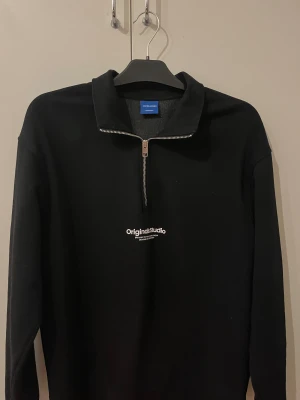 Svart half zip tröja Jack & Jones - Svart långärmad tröja från Jack & Jones med halv dragkedja och tryckt text 'Original Studio' på bröstet.Materialet är mjukt 