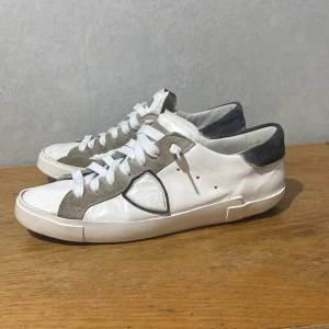Philippe Model vita sneakers i skinn - Snygga vita sneakers från Philippe Model med grå mockadetaljer och mörkgrå häl. Skorna har klassisk snörning, rund tå och platt sula. Tillverkade i skinn och mocka för en lyxig känsla. Perfekta för dig som gillar stilrena och trendiga sneakers. Hål under skorna, kmr in vatten i därför säljer jag billigt.