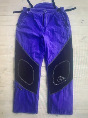 90s Quiksilver Snowboardbyxor Vintage - Unika och riktigt retro snowboardbyxor från tidigt 1990-tal, original från Quiksilver. Byxorna är i otroligt fint skick och har mest legat nedpackade i garderoben – perfekt för dig som älskar äkta 90-talsstil eller vill komplettera ett vintage set.  💜 Färg: Lila med svarta paneler 📏 Storlek: Large 🎿 Detaljer: Klassiska förstärkningar, justerbara remmar och den ikoniska 90’s designen ✨ Skick: Nyskick för sin ålder  Vintage snowboardkläder från denna era är mycket eftertraktade – extra roligt i en
