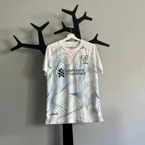 Liverpool Salah 11 vit fotbollströja - Snygg vit Liverpool fotbollströja med M. Salah och nummer 11 på ryggen. Framsidan har klubbmärke och sponsortryck samt vågiga linjer i blått, grönt och rött. Kortärmad och tillverkad i lätt polyester som andas. Perfekt för dig som älskar fotboll och Salah.