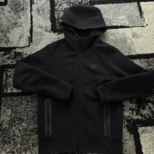 Svart Nike tech - Svart Nike tech från Nike med hel dragkedja framtill och två fickor med dragkedja. Hoodien har en stilren look och är tillverkad i mjukt, stretchigt material. Perfekt för chill dagar och kalla.