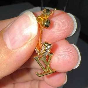 Luis vytton örhängen i guld med stenar - Snygga örhängen i guldton med logga och glittrande stenar runt hela ringen. Perfekt accessoar för dig som vill sticka ut och addera lite lyx till din stil. Klassisk hoop-modell med exklusiv känsla och detaljer som verkligen syns.