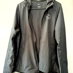 Svart Nike Air vindjacka med huva - Svart Nike Air vindjacka med huva och dragkedja framtill. Jackan har logga på bröstet och stor Nike Air-tryck på ryggen. Lätt material, perfekt för blåsiga dagar. Två sidofickor och justerbar huva med snörning. Stilren och sportig look.