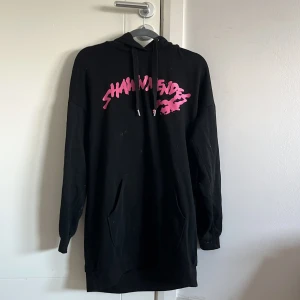 Svart hoodieklänning Shawn Mendes - Svart oversized hoodieklänning från Shawn Mendes med rosa tryck fram och bak. Stor känguruficka, huva med snörning och långa ärmar. Perfekt för dig som gillar streetstyle och vill ha något bekvämt men ändå snyggt.