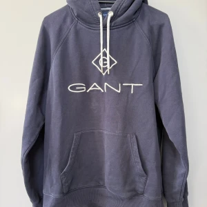 Mörkblå hoodie från GANT med logga - Säljer en mörkblå hoodie från GANT med stor vit broderad logga på bröstet. Tröjan har huva med vita snören och en stor magficka framtill. Perfekt för dig som gillar klassisk streetstil och vill ha något bekvämt och snyggt.
