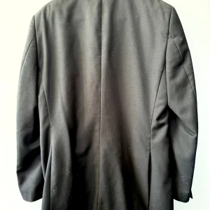 Svart klassisk kavaj med två knappar - En svart kavaj med två knappar framtill, klassisk krage och raka fickor. Kavajen har en stilren och tidlös design, perfekt för dig som vill ha en clean och enkel look. Materialet känns slätt och fodret är också svart. Passar till många olika stilar.