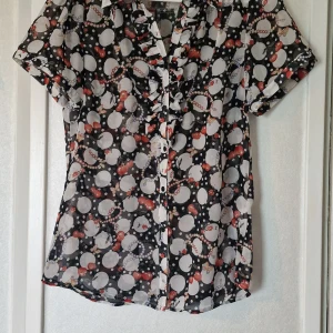 Mönstrad kortärmad blus från Red Label - Snygg blus från Red Label i storlek L med svart botten och stora vita prickar, röda detaljer och pärlmotiv. Blusen har korta ärmar, volang vid knäppningen och är tillverkad i ett tunt, lätt material som känns luftigt. Dtorlekdmärkningen är dock något skev. Jag skulle snarare säga att den storleksmässigt är som en S eller M.