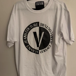 Vit t-shirt från Versace Jeans Couture - Snygg vit t-shirt från Versace Jeans Couture med stort svart tryck framtill. Klassisk passform och rund halsringning. Perfekt för dig som vill ha en stilren och trendig look med tydlig logga. Gjord i mjuk bomull som känns skön mot huden. Storlek L, använd 1 gång så i princip ny.