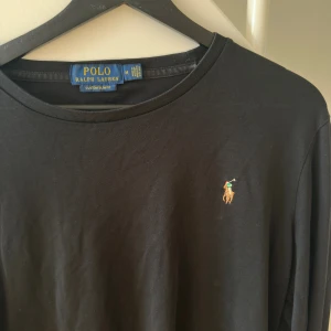 Svart långärmad tröja Polo Ralph Lauren - Svart långärmad tröja från Polo Ralph Lauren i slim fit-modell. Klassisk design med rund halsringning och den ikoniska broderade polospelaren på bröstet. Tillverkad i mjuk bomull som känns skön mot huden. Perfekt för en clean och stilren look.