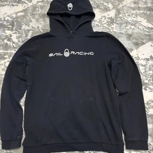 Svart hoodie från Sail Racing - Svart hoodie från Sail Racing med vit logga på bröstet och tryck på huvan. Klassisk känguruficka framtill och mjuk bomullskänsla. Perfekt för dig som gillar en clean och sportig stil.