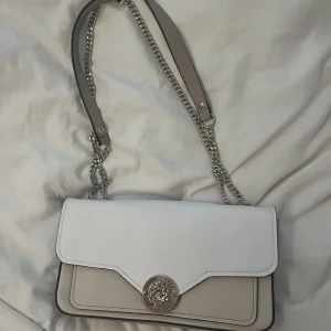 Vit och beige axelväska från Guess - Snygg axelväska från Guess i vitt och beige med silverfärgad kedja och läderdetaljer. Väskan har ett stilrent metallspänne framtill med Guess-logga och en elegant, strukturerad design.
