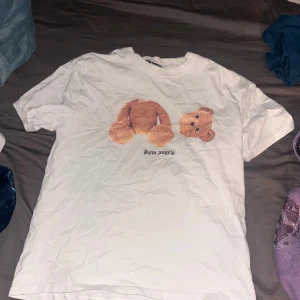 Vit Palm Angels t-shirt med björntryck - Säljer en vit t-shirt från Palm Angels med ikoniskt björntryck framtill och stor logga på ryggen. Klassisk passform och rund hals. Perfekt statement-plagg för dig som gillar streetwear och vill sticka ut.