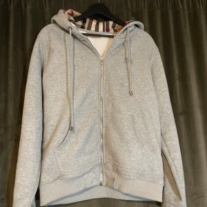 Grå hoodie från Burberry med dragkedja - Grå hoodie från Burberry i storlek L med klassiskt rutigt foder i beige, svart och rött. Tröjan har huva med snörning, dragkedja framtill och två fickor. Perfekt för dig som vill ha en stilren och lyxig vardagslook.
