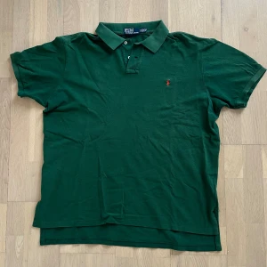 Grön pikétröja från Ralph Lauren - Snygg grön pikétröja från Ralph Lauren med klassisk krage och två knappar framtill. Tröjan har korta ärmar och är tillverkad i mjuk bomull. På bröstet finns det ikoniska röda Polo-logotypen broderad. Perfekt för en chill och stilren look.