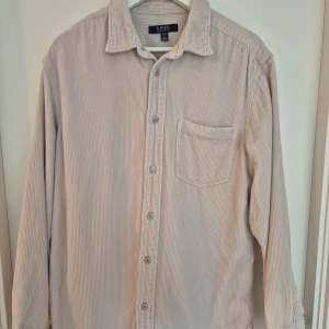 Beige manchesterskjorta från SMOG - Säljer en beige manchesterskjorta från SMOG i regular fit. Skjortan har klassisk krage, bröstficka och knäppning framtill. Materialet är mjukt och ger en chill vibe, perfekt för lager-på-lager. Långärmad och med diskreta ränder i tyget.