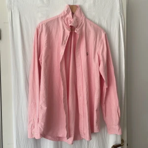Rosa skjorta från Polo Ralph Lauren - Säljer en klassisk rosa skjorta från Polo Ralph Lauren i bomull. Skjortan har button-down krage, broderad logga på bröstet och långa ärmar. Passformen är normal och modellen är Classic Fit. Perfekt för dig som gillar stilrena och tidlösa plagg.
