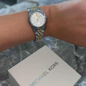 Michael Kors Lexington klocka silver/guld - Säljer en snygg Michael Kors Lexington klocka i silver och guld med klassisk länkarmband. Urtavlan är vit med gulddetaljer och små stenar som markörer. Klockan har en stilren och tidlös design som passar till många olika looks. Kan gå ner i pris vid snabb affär, skriv för intresse🩷
