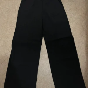 Svarta vida byxor från H&M - Svarta byxor från H&M med breda ben och resår i midjan. De har en enkel och stilren look, perfekta att matcha med en crop top eller hoodie. Materialet känns lätt och bekvämt, och passformen är loose och relaxed.