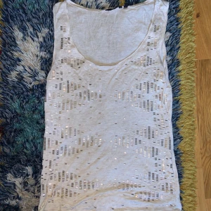 Vit linne med paljettmönster - Säljer ett vit linne med snyggta paljetter i guld och rund halsringning. Toppen är lätt och luftig i mjukt material.