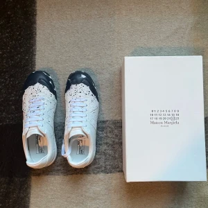 Maison Margiela paint splatter - Unika vita sneakers från Maison Margiela med svart färgstänk och svart tåparti. Skorna har snörning, rund tå och är tillverkade i skinn. Snygg och konstnärlig design som sticker ut, levereras med originalkartong.