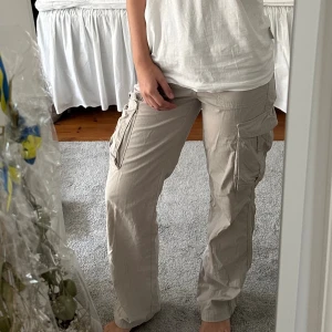 Beige cargo straight byxor från Bershka - Säljer ett par beigea cargo straight byxor från Bershka i storlek 34. Byxorna har stora fickor på sidorna samt på rumpan med knappar, raka ben och justerbara spännen i midjan. Tillverkade i bomull för en skön och avslappnad look. Perfekta för en chill och trendig stil.