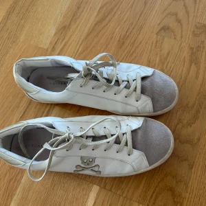 Vita Scalpers sneakers med dödskalle - Snygga vita sneakers från Scalpers med grå mockadetalj på tån och cool dödskalle-logga på sidan. Skorna har snörning och platt sula, samt diskreta gulddetaljer på hälen. Perfekta för dig som gillar en edgy och stilren look. Använda 4 ,5 gånger. Finare  i verkligheten 