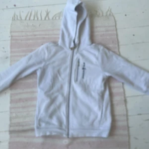 Vit hoodie från Sail Racing - Vit hoodie från Sail Racing med dragkedja framtill och broderad logga på bröstet. Tröjan har huva, långa ärmar och en praktisk bröstficka med dragkedja. Tillverkad i mjuk bomullsmix, perfekt för chill dagar. Använt ungefär 2 gånger, i bra skick. Köpte för 800
