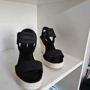 Svarta kilklack-sandaler från DKNY - Snygga svarta sandaler från DKNY med breda band och hög kilklack i flätad beige textil. Skorna har öppen tå och plattformsula, perfekt för sommarens alla outfits. Guld-detalj på hälen ger extra lyxig känsla.