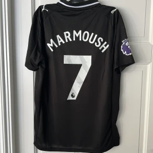 Manchester City svart fotbollströja Puma - Snygg svart Manchester City fotbollströja från Puma med Marmoush och nummer 7 på ryggen. Tröjan har korta ärmar, broderad klubbmärke, Premier League-patch på ärmen och Etihad Airways-tryck i silver. Tillverkad i lätt och ventilerande funktionsmaterial. Lite prickar vid sponsorn. Replika. Endast handtvätt gäller!