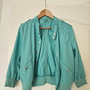 Turkos bomberjacka med silverknappar - Snygg turkos bomberjacka med ribbade muddar och silverfärgade knappar. Jackan har dragkedja framtill, två fickor med tryckknappar och coola axeldetaljer. Perfekt för dig som vill sticka ut med färg i vardagen.