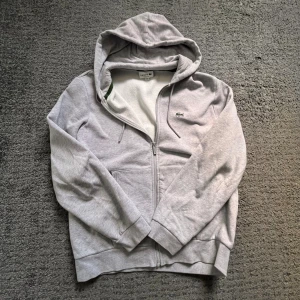Grå hoodie från Lacoste med dragkedja - Snygg ljusgrå hoodie från Lacoste i klassisk passform. Tröjan har huva med snörning, dragkedja framtill och två fickor. Ikoniska Lacoste-loggan på bröstet. Perfekt för kalla höst och vinterdagar!